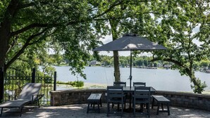 Une terrasse avec une table et des chaises, un grand parasol et une vue sur un lac où naviguent des bateaux.
