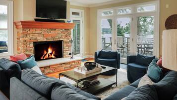 A living room with a stone fireplace, a flat-screen TV mounted above it, and a large window overlooking a deck.