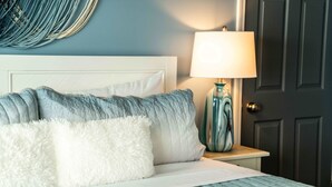 Un lit bien fait, avec une tête de lit blanche, une couvre-lit à rayures bleues et blanches, et de moelleux oreillers blancs. Une lampe turquoise avec un abat-jour blanc est posée sur une table de chevet à côté du lit.