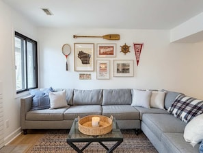 Uma sala de estar com um sofá cinza, uma mesa de centro de vidro e uma parede decorada com quadros e uma prancha.