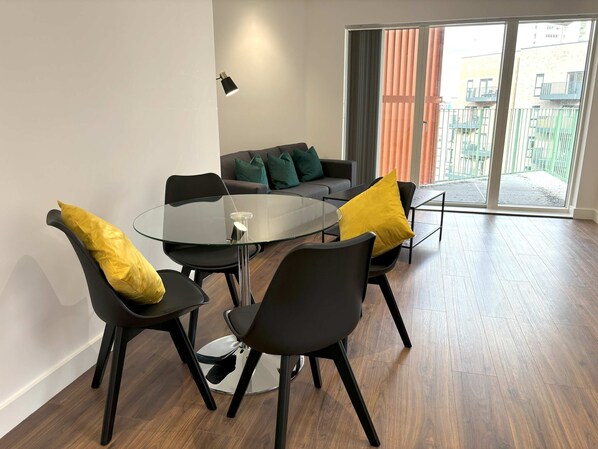 Un salón moderno con una mesa de vidrio rodeada de sillas negras con cojines amarillos, un sofá gris con cojines verdes y un amplio ventanal corredero que da a un espacio exterior.