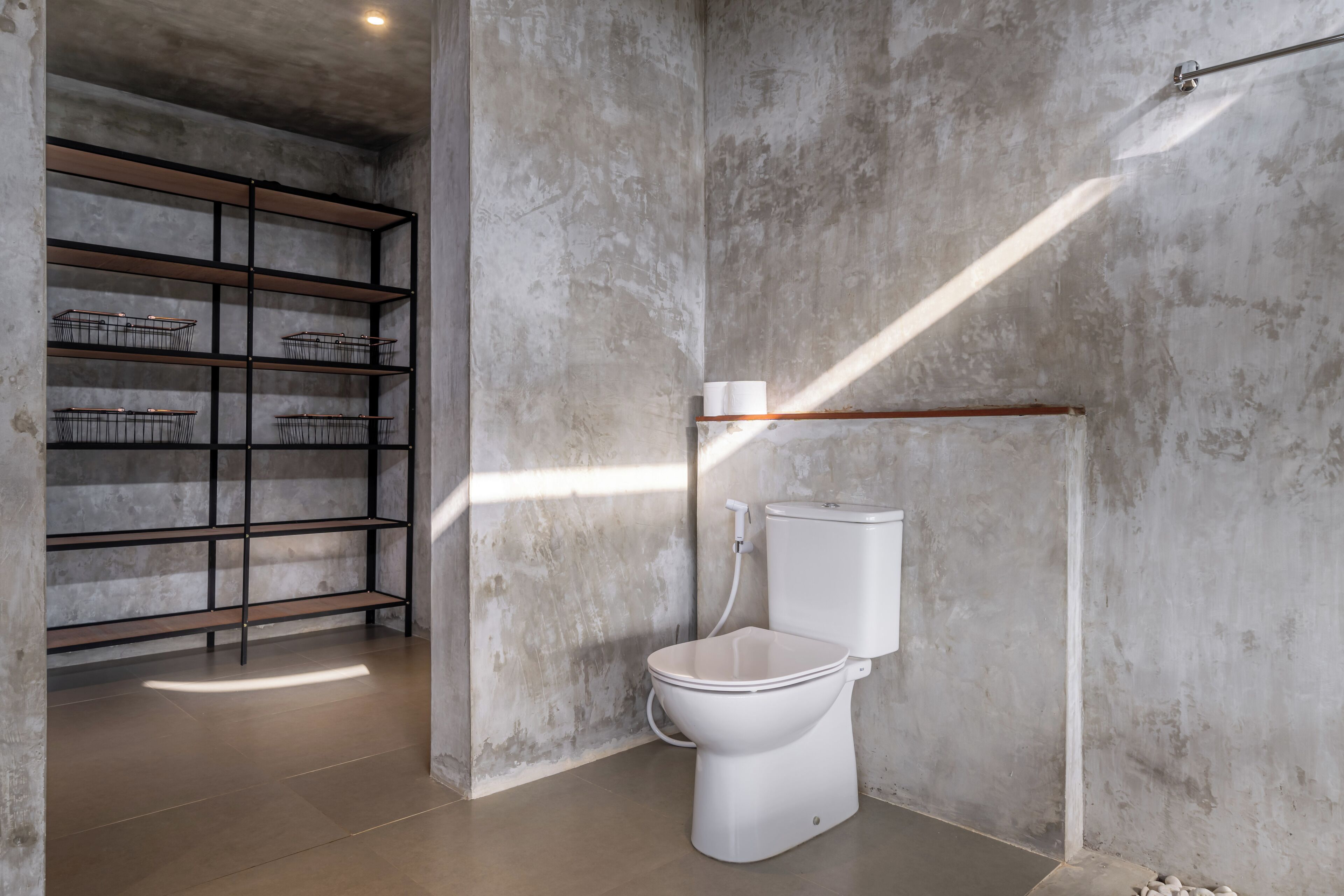Un baño con inodoro blanco, una estantería metálica y pared de concreto.
