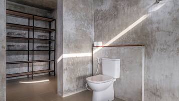 Un baño con inodoro blanco, una estantería metálica y pared de concreto.