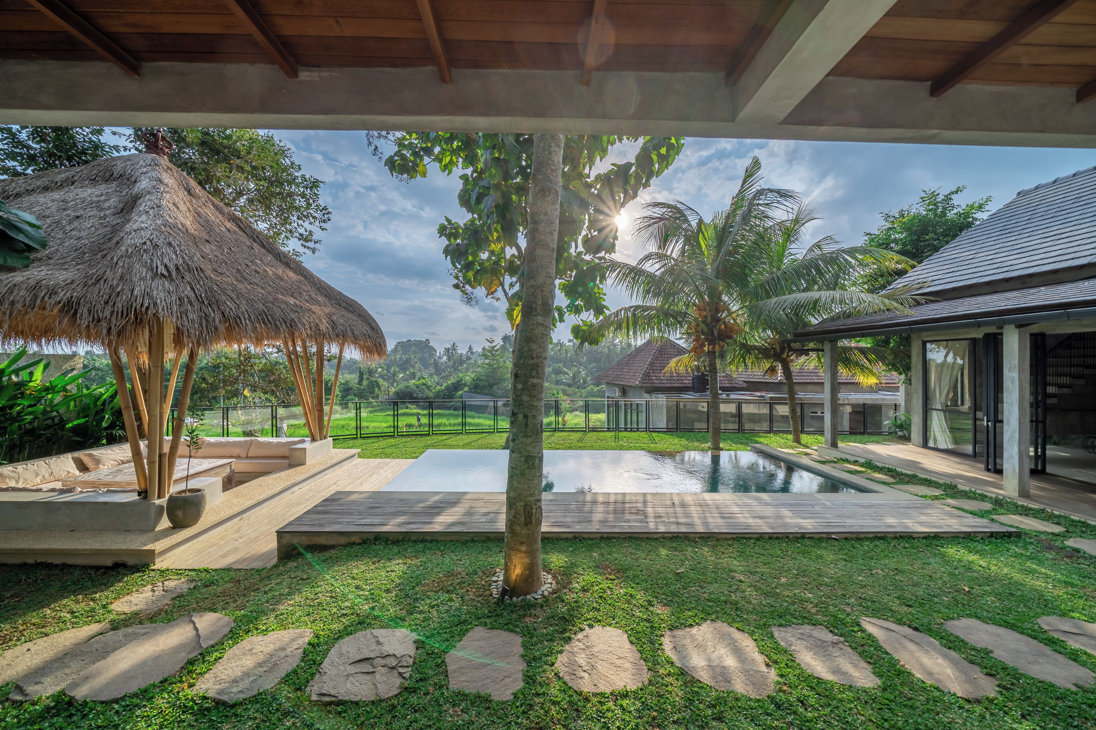 A modern outdoor area with a pool, stone pathways, a thatched roof structure, and a view of a residential area.
