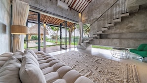 A spacious living area with a large sofa, a patterned rug, a green chair, and a staircase leading to an outdoor area. - Sola Villa Ubud - 2 Bedrooms (Ubud)