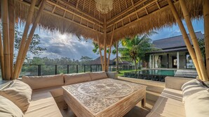A thatched-roof outdoor seating area with a pool view, a large ornate coffee table, and a modern building in the background. - Sola Villa Ubud - 2 Bedrooms (Ubud)