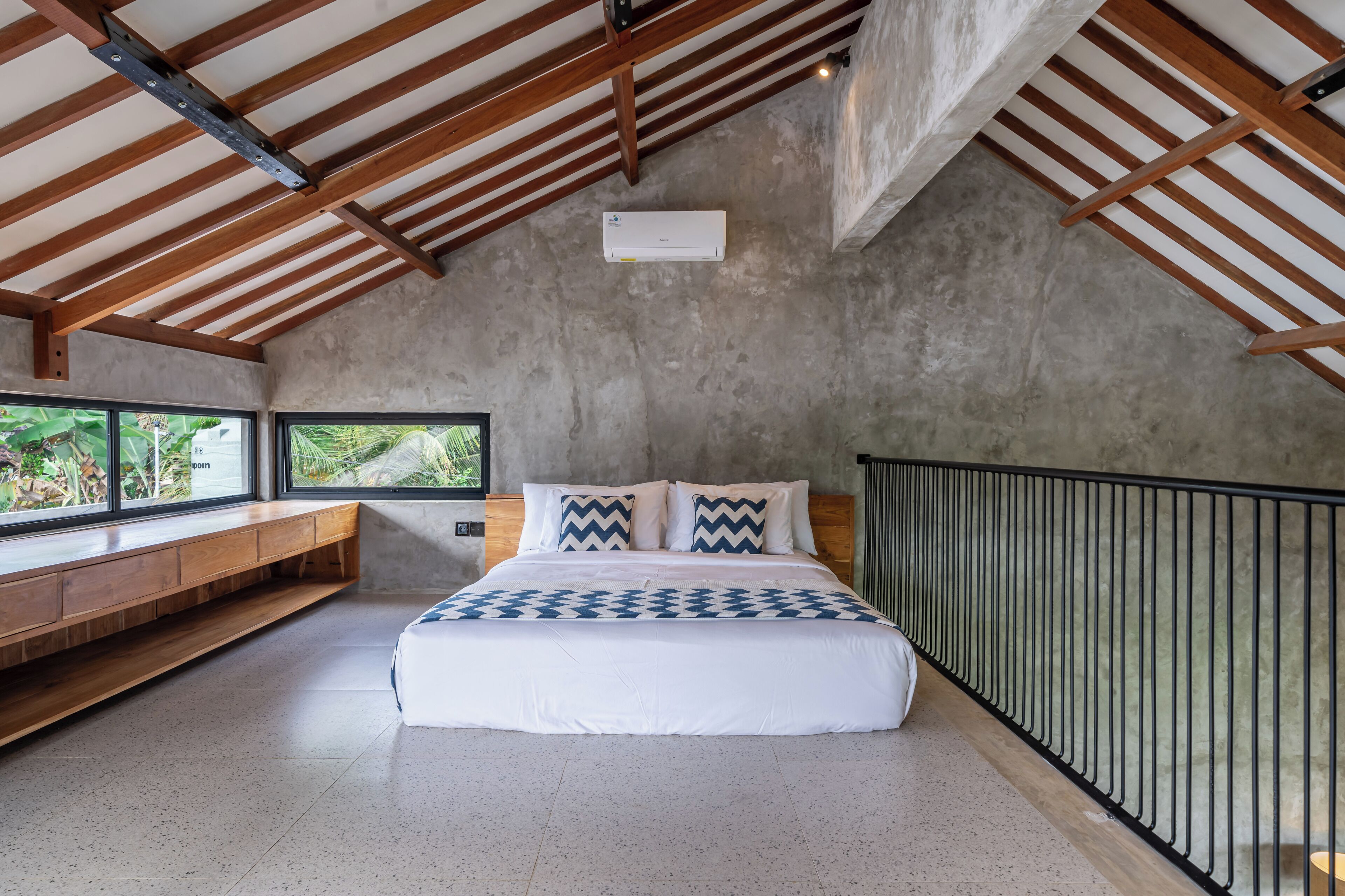 Een slaapkamer met een groot bed, houten balken, een betonwand, een raam en een balkongevel.