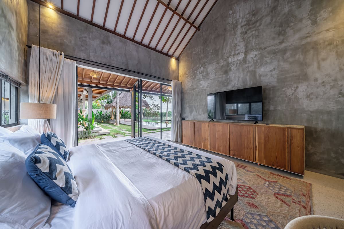 Une chambre moderne avec un grand lit, un téléviseur posé sur un meuble en bois et une vue sur un jardin grâce à de grandes fenêtres.