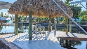 A thatched-roof outdoor seating area with a pool in the background. - Serenity by the Gulf - Vesteva Vacation Rentals (Cape Coral)