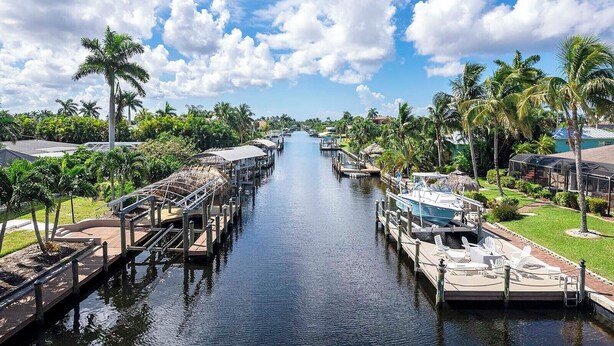 A canal with boats docked on both sides, surrounded by palm trees and buildings with thatched roofs. - Serenity by the Gulf - Vesteva Vacation Rentals (Cape Coral)