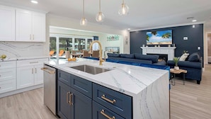 A modern kitchen with a marble island, dark cabinetry, and gold handles. There is a dining area with a blue sofa and a fireplace. - Serenity by the Gulf - Vesteva Vacation Rentals (Cape Coral)