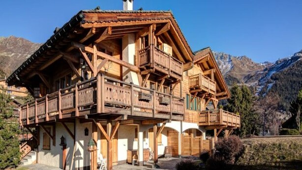 Eine Holzhütte mit mehreren Balkonen, einem Steinfundament und einem Bergpanorama im Hintergrund.