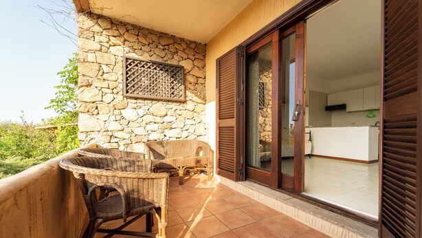 A balcony with wicker furniture, a stone wall, and a sliding glass door leading to a room.