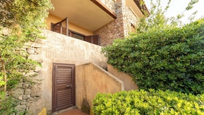 A narrow courtyard with a stone wall, a wooden door, and lush green bushes.