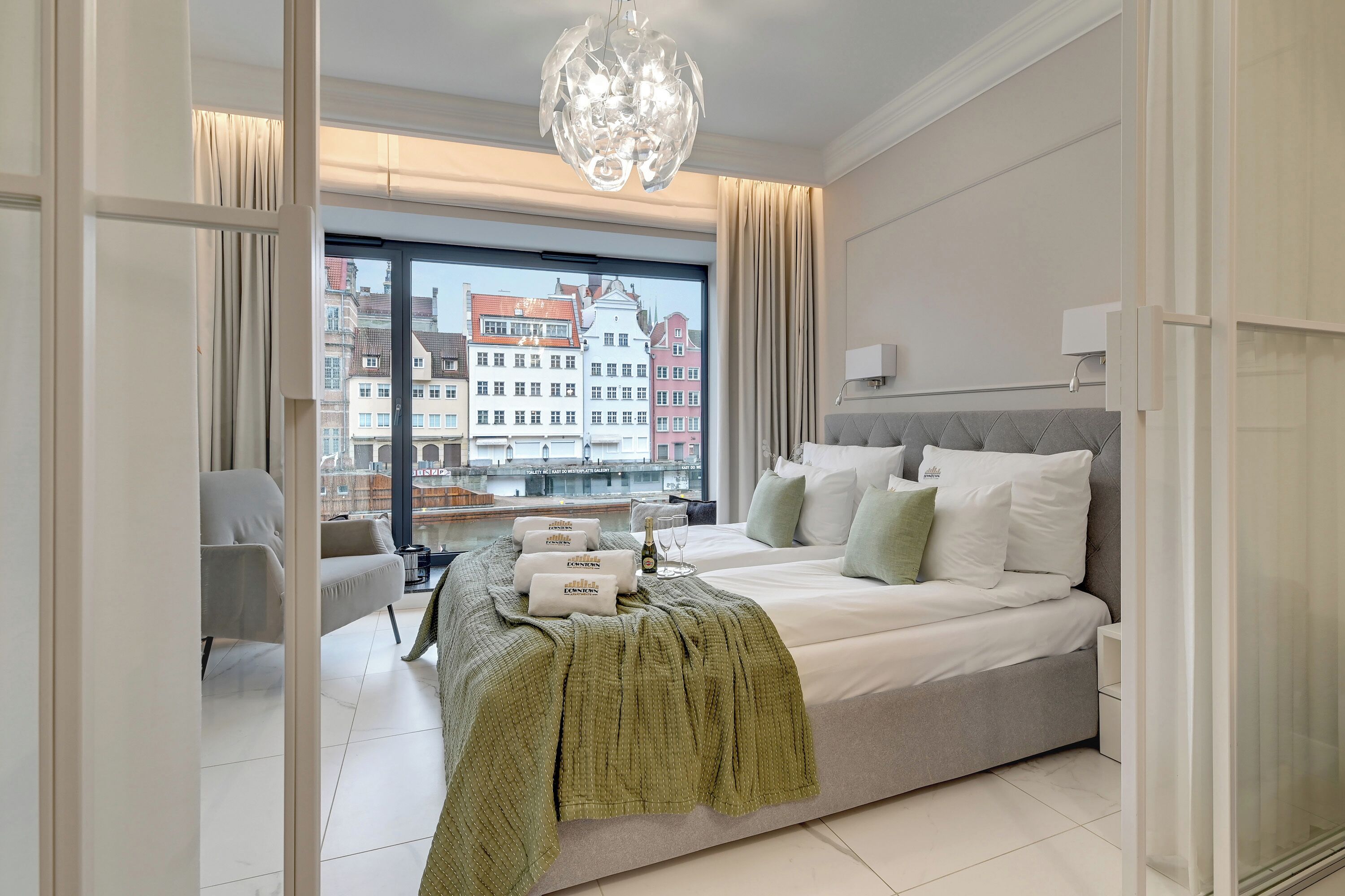 A modern bedroom with a large bed, a view of buildings through a window, and a chandelier.