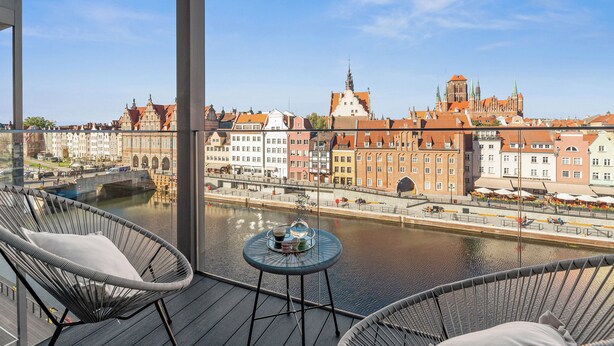A balcony with wicker chairs and a small table, overlooking a river with historic buildings and a bridge. - Deo Plaza by Downtown Apartments (Gdansk)