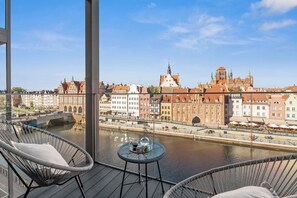 Varanda com cadeiras de vime e uma mesa pequena, com vista para um rio e edifícios históricos e uma ponte.