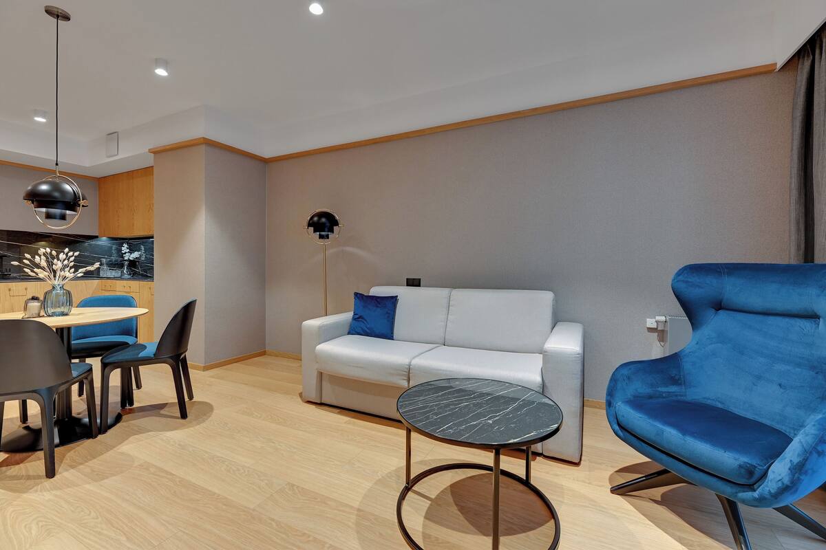 A modern living room with a white sofa, blue armchair, round table, and a dining area with chairs and a vase.
