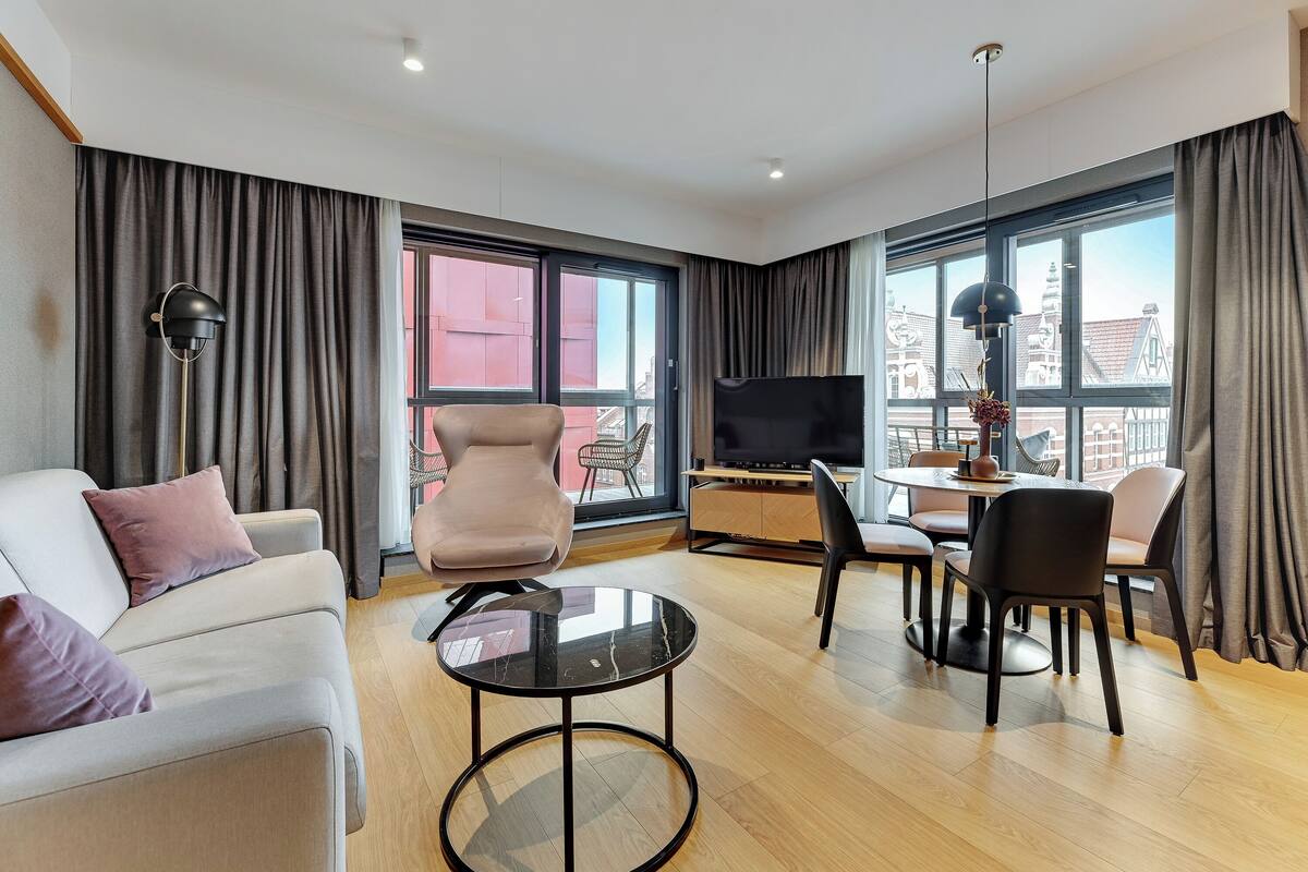 A modern hotel room with a sofa, a dining table with chairs, a TV, and large windows with curtains.