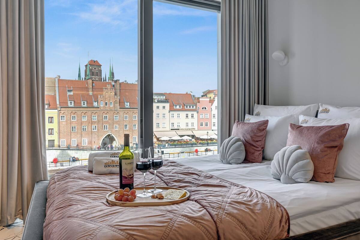 A hotel room with a large window offering a view of historic buildings and a river. The bed is neatly made with a pink blanket and decorative pillows.