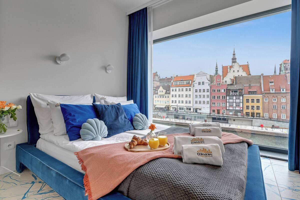 A modern hotel room with a large bed, a view of a cityscape, and breakfast items on the bed.