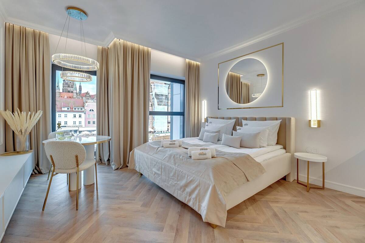 A modern hotel room with a large bed, a round mirror, a dining table with chairs, and a view of the city through large windows.