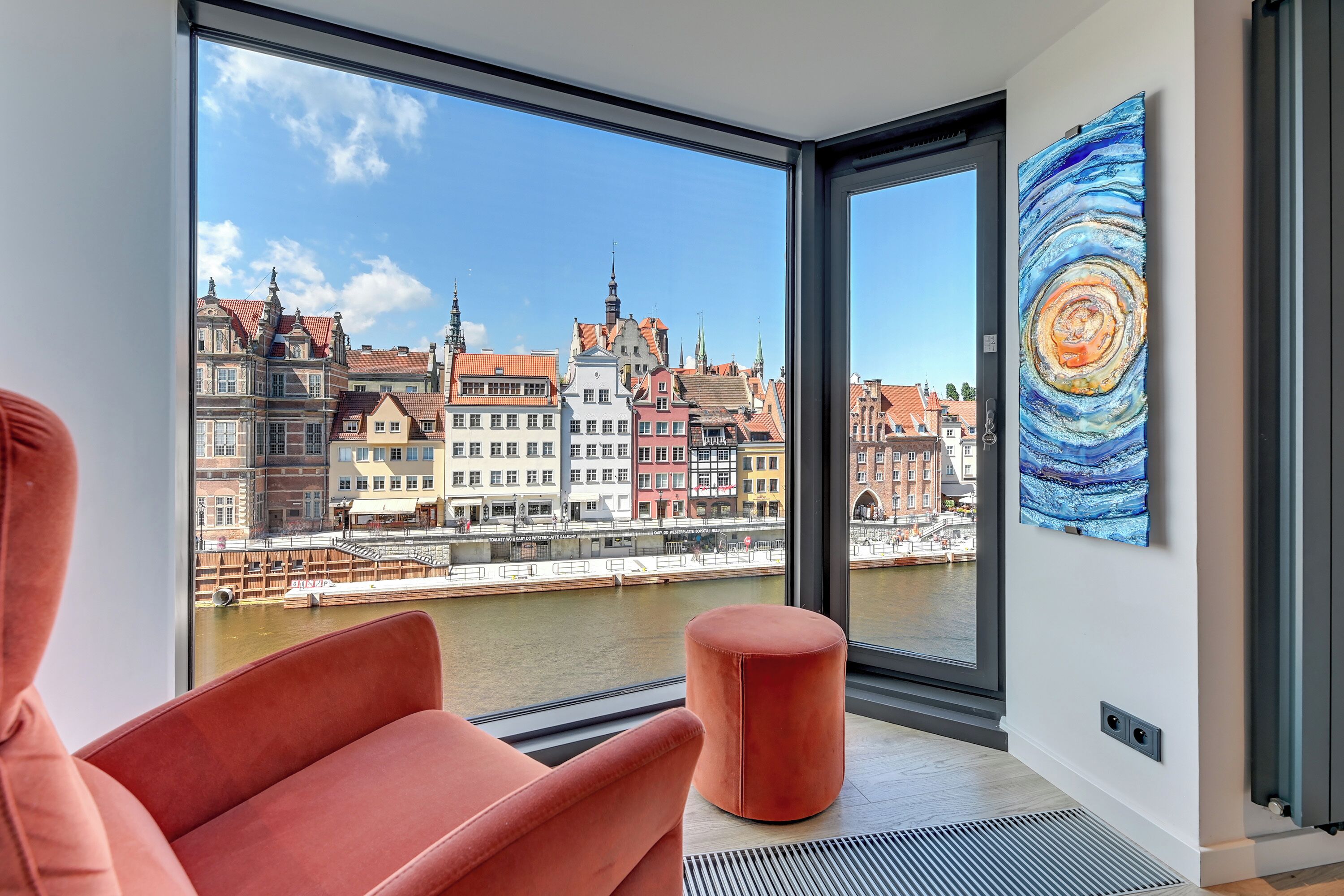 Ein modernes Zimmer mit großem Fenster, einem roten Sessel, einem kleinen Hocker und einem farbenfrohen Gemälde an der Wand.