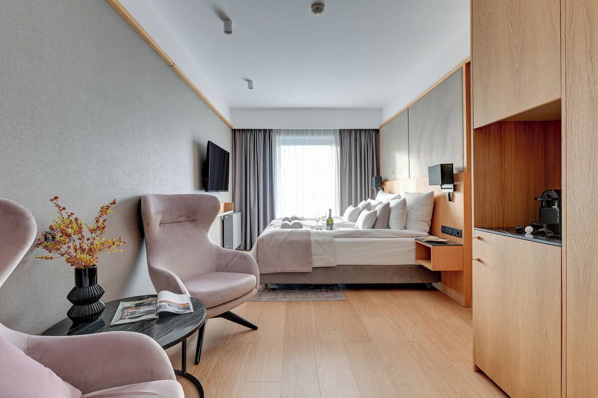 A modern hotel room with a bed, two chairs, a small table with a vase, a flat-screen TV, and a wooden cabinet.
