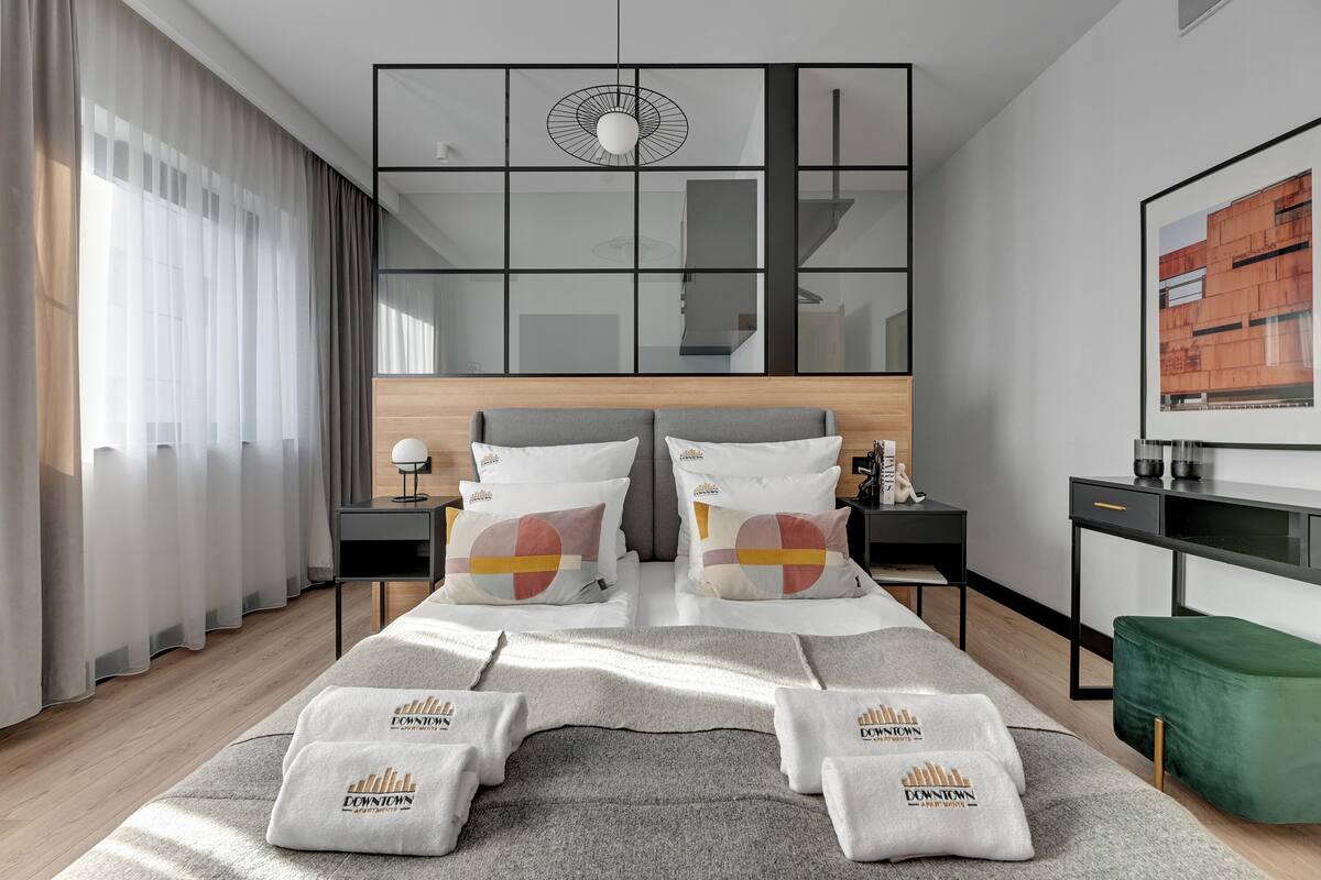 A modern hotel room with a large bed, a desk, a green ottoman, and a large window with sheer curtains.