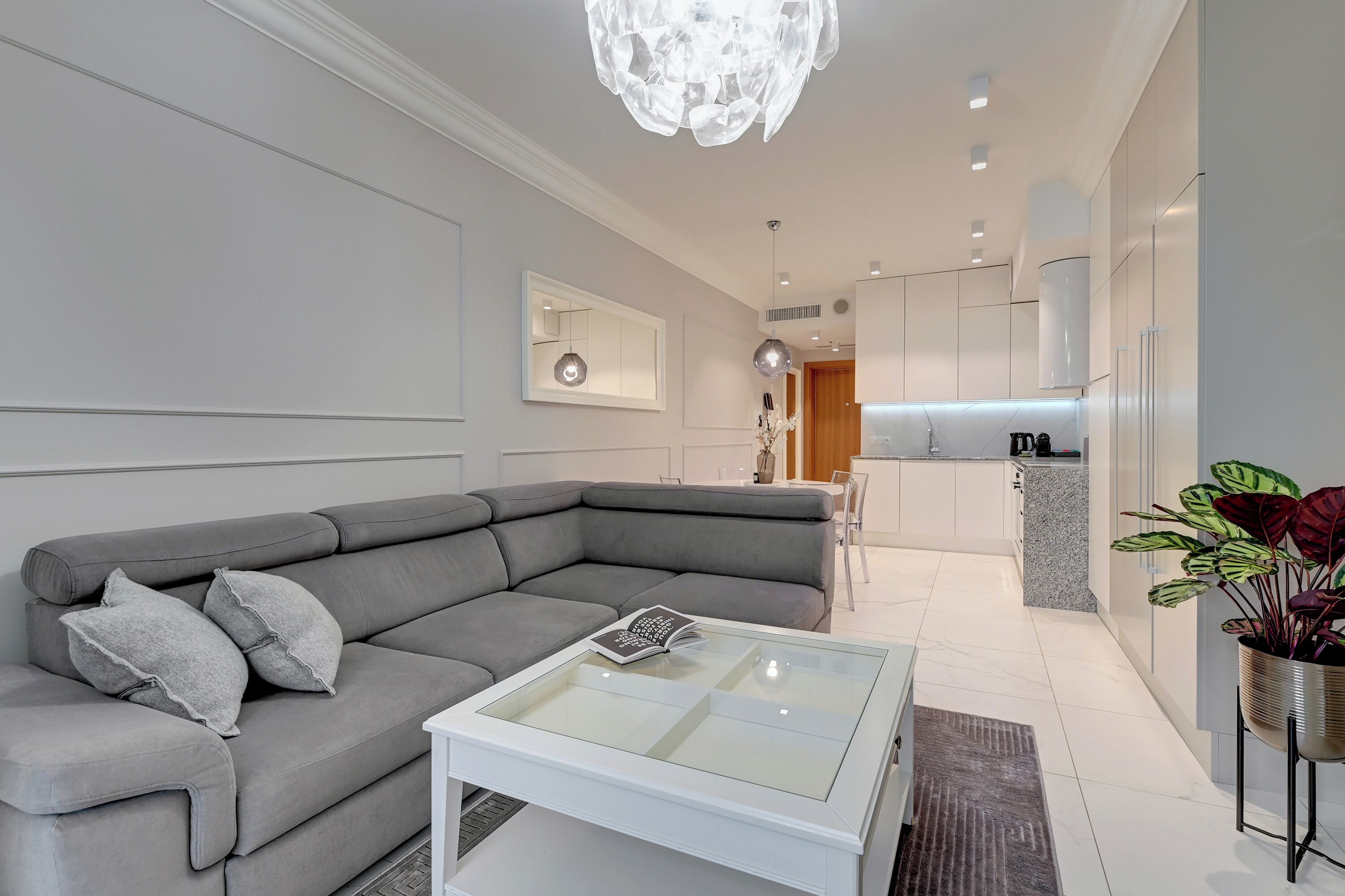 A modern living room with a grey sectional sofa, a glass coffee table, and a kitchen in the background.