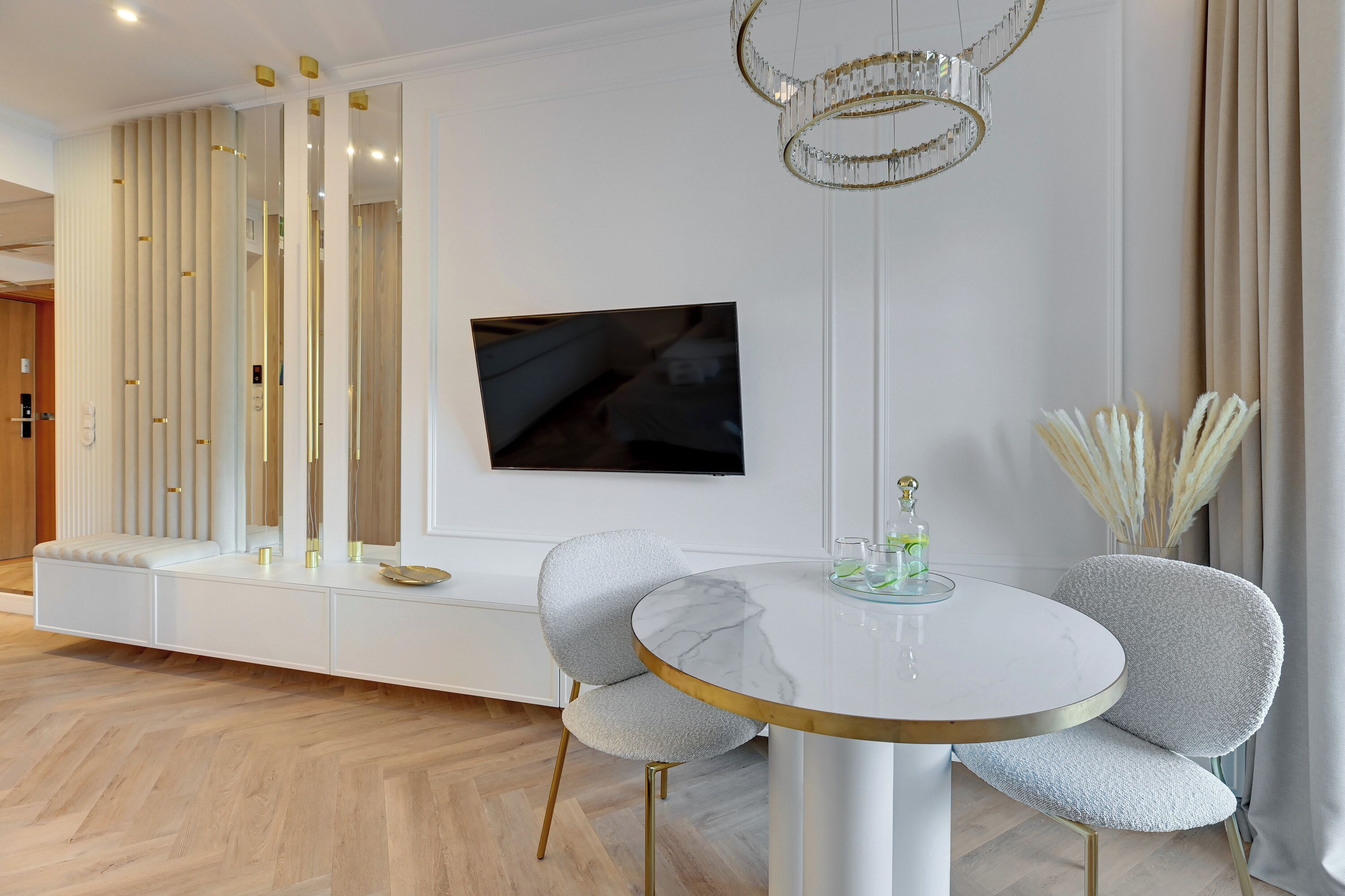 A modern dining area with a round table, two grey upholstered chairs, a white console table, a flat-screen TV mounted on the wall, and a decorative chandelier.