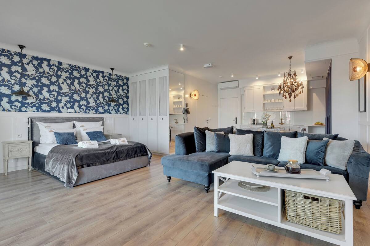 A modern bedroom with a bed, a sofa, a coffee table, and a kitchen area visible through an open door.