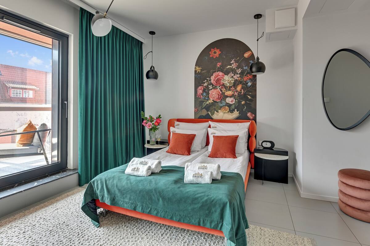 A modern bedroom with a large bed, green curtains, a floral painting, a round mirror, and a view of the outside.