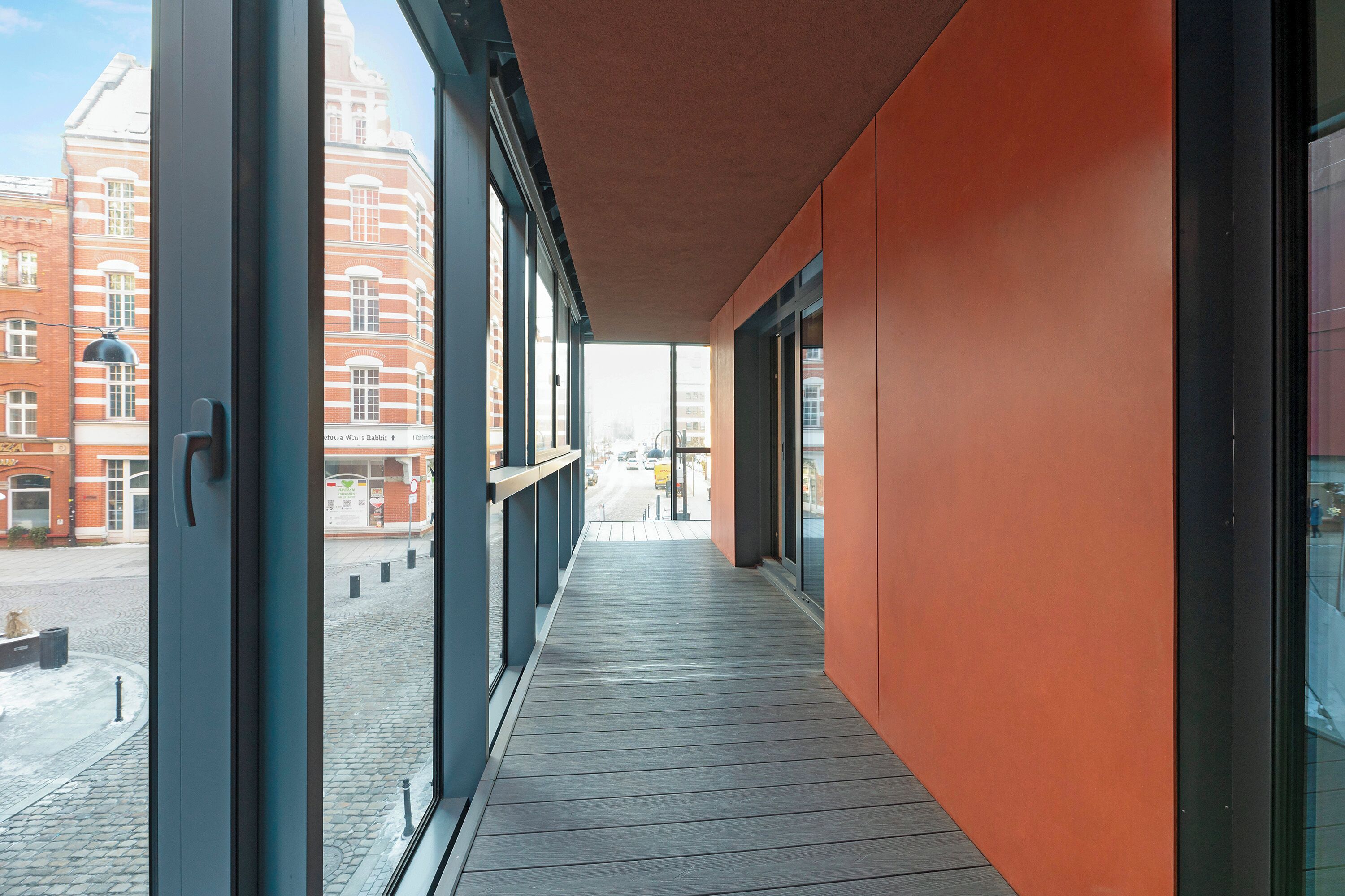 Un pasillo con amplios ventanales, suelo de madera y vista a una calle con edificios.