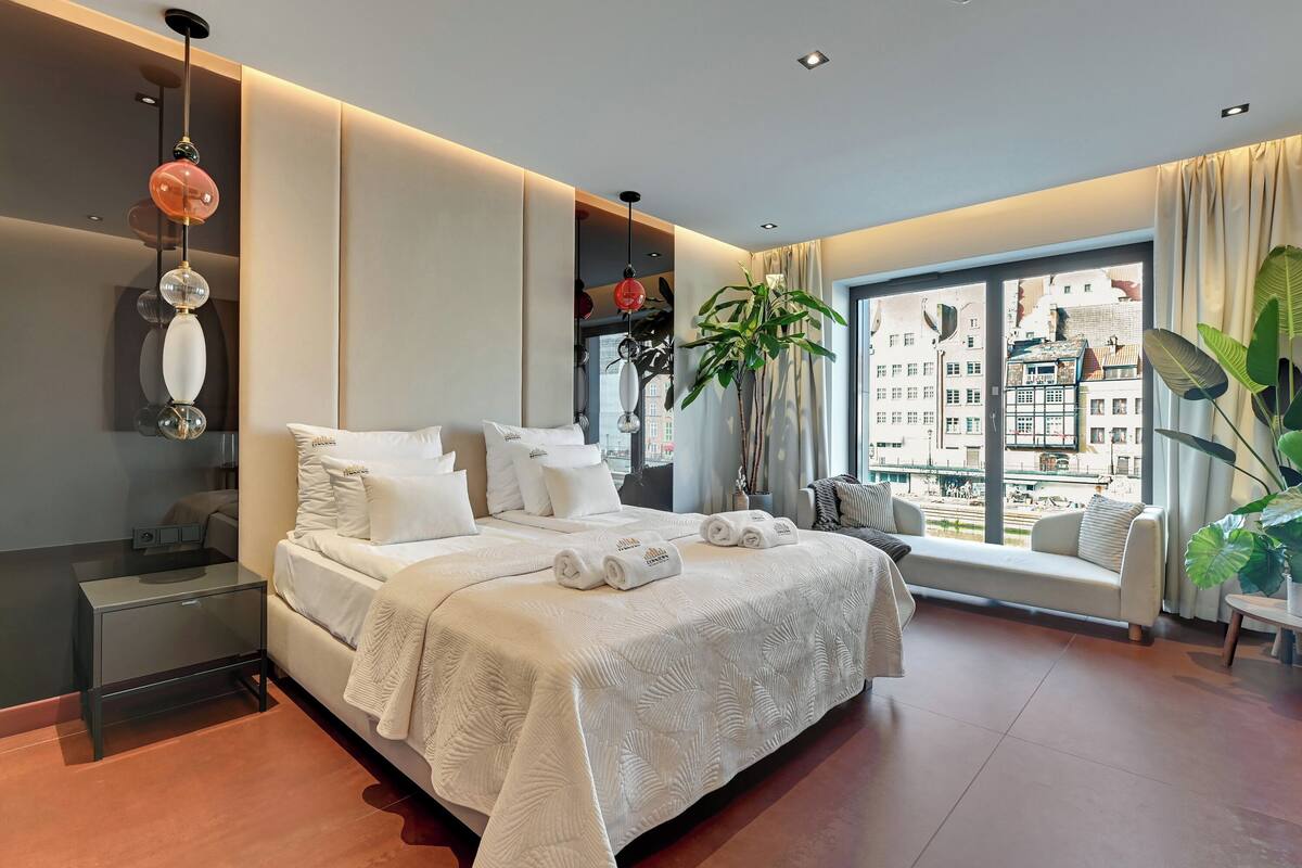 A modern hotel bedroom with a large bed, a bedside table, a window with a city view, and a potted plant.