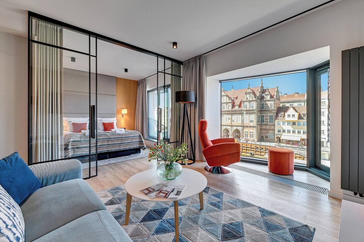 A modern hotel room with a large window, a bed, a sofa, a coffee table, and a red chair.