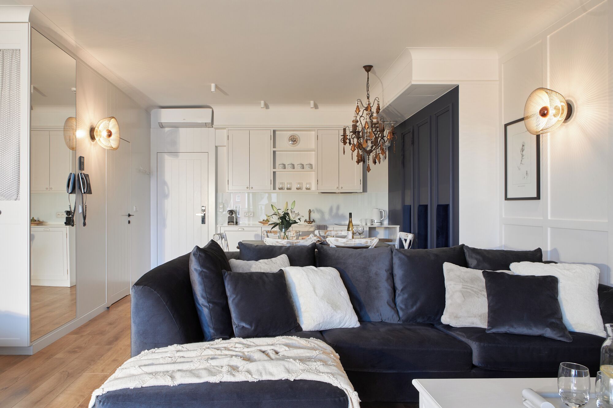 Uma sala de estar moderna com um sofá azul escuro, um cobertor branco e um candelabro. Uma cozinha com armários brancos e uma área de jantar com mesa e cadeiras são visíveis ao fundo.