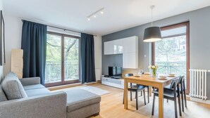 A modern living room with a grey sofa, a dining table with chairs, a TV, and a large window with curtains. - Imperial Apartments - Mewa (Sopot)