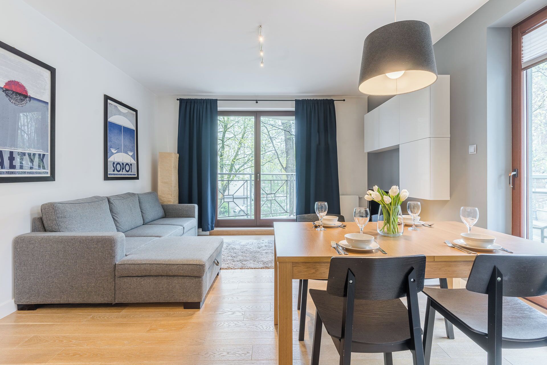 Una sala de estar moderna con un sofá gris, una mesa de comedor lista para cuatro comensales y un gran ventanal con cortinas.