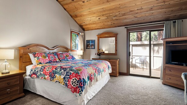 Un dormitorio con techo de madera, cama con colcha floral de colores, mesita de noche con lámpara, cómoda y televisor.