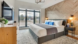 A modern bedroom with a large bed, a wooden dresser, a TV, and a view of the outdoors. - Mountain-view Condo in Downtown Winter Park! (Winter Park)