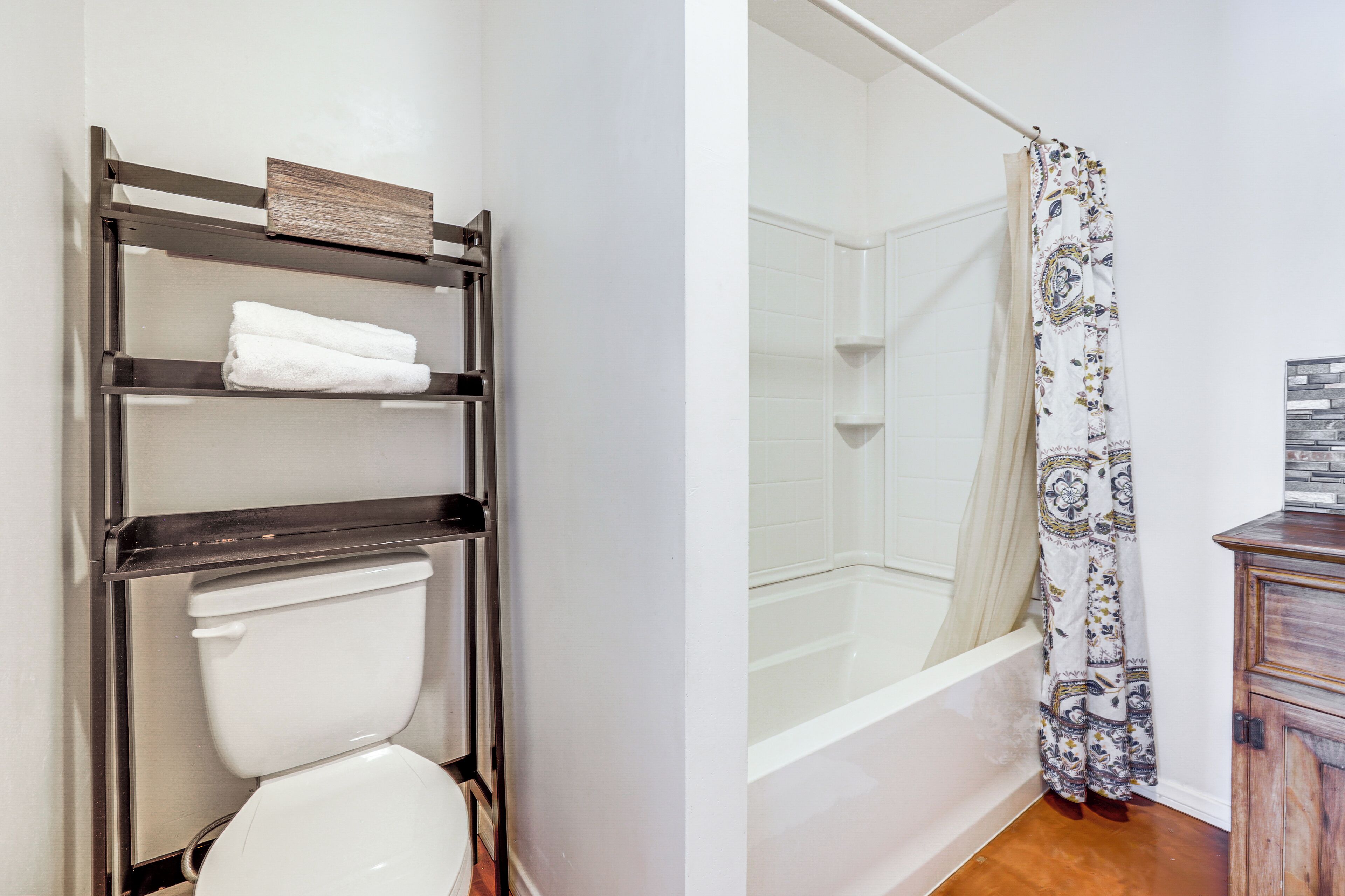 A bathroom with a toilet, a shelf with towels, a shower with a patterned curtain, and a wooden cabinet.