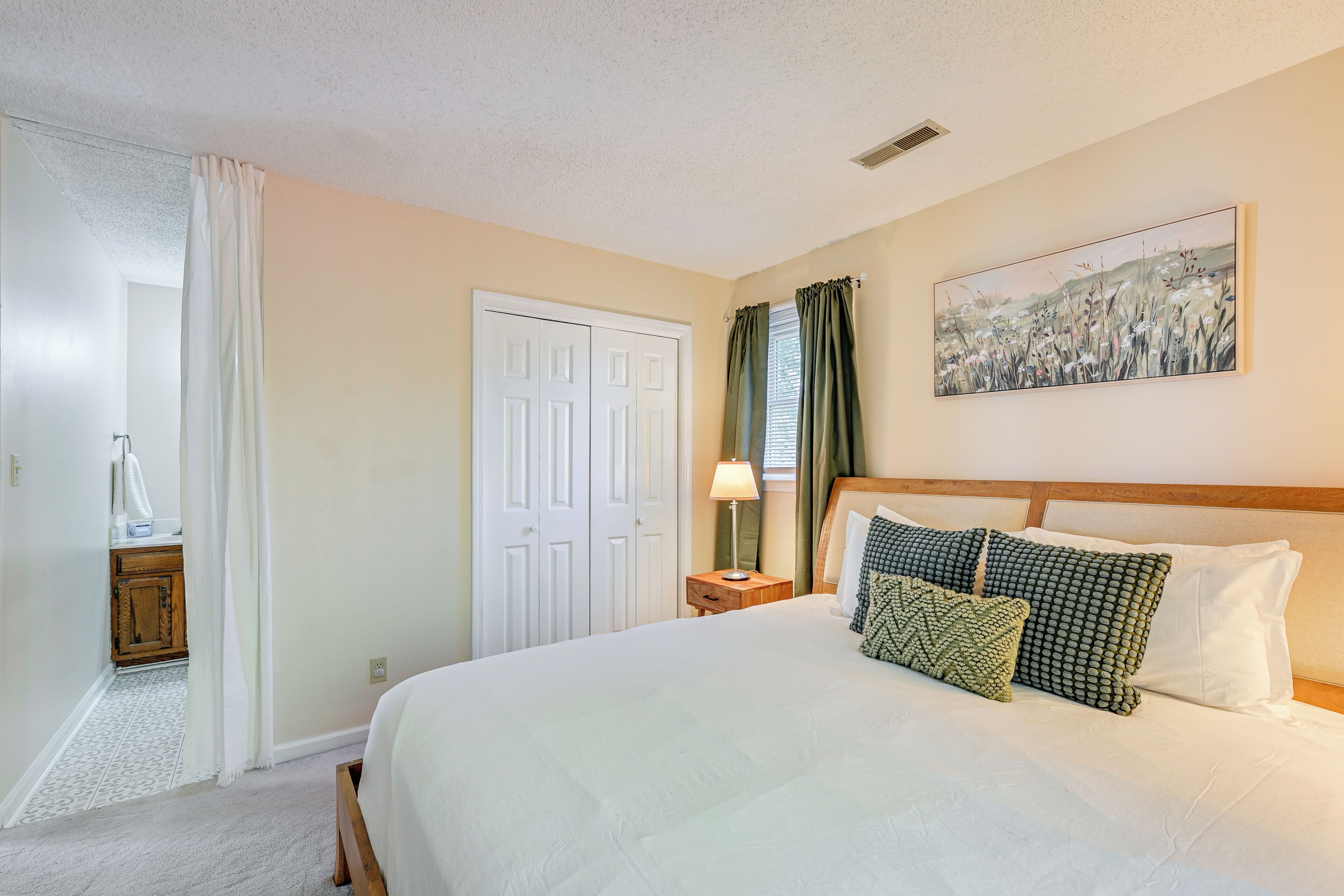 A bedroom with a large bed, a nightstand with a lamp, a door, and a painting on the wall.