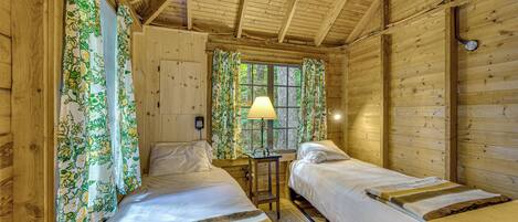 A wooden cabin room with two beds, a table, and a window with curtains.