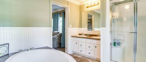 A bathroom with a large bathtub, a shower, a vanity with a mirror, and a door leading to another room.