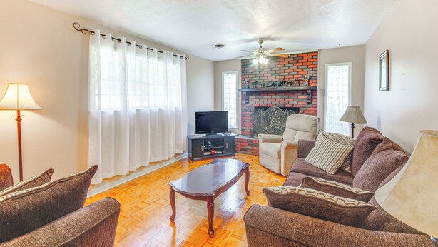 A living room with a wooden floor, a television, a fireplace with a brick wall, and a ceiling fan.