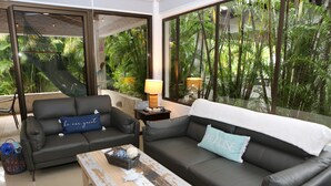 Ein modernes Wohnzimmer mit zwei schwarzen Ledersofas, einem Holztisch und großen Fenstern mit Blick auf üppiges Grün.