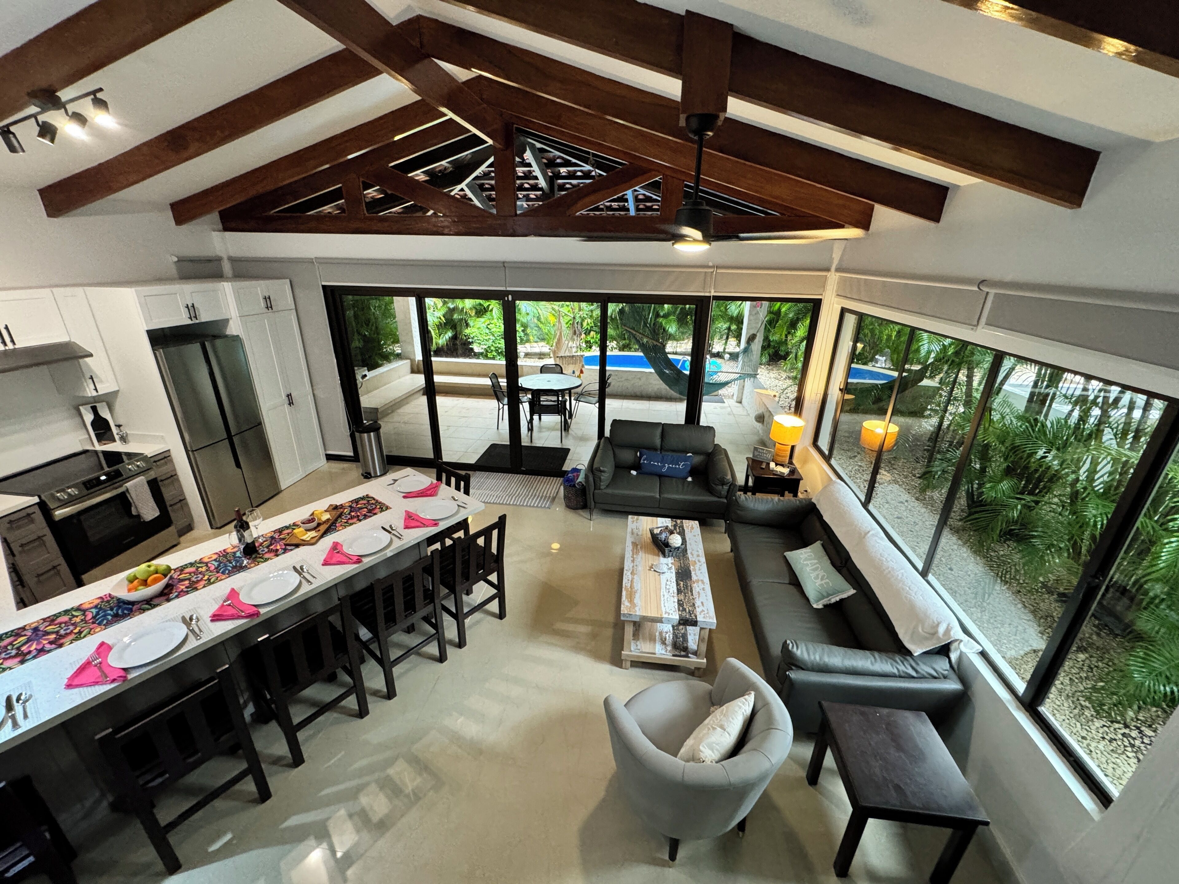 A modern kitchen with a dining area, a sofa set, and a view of a swimming pool and greenery.