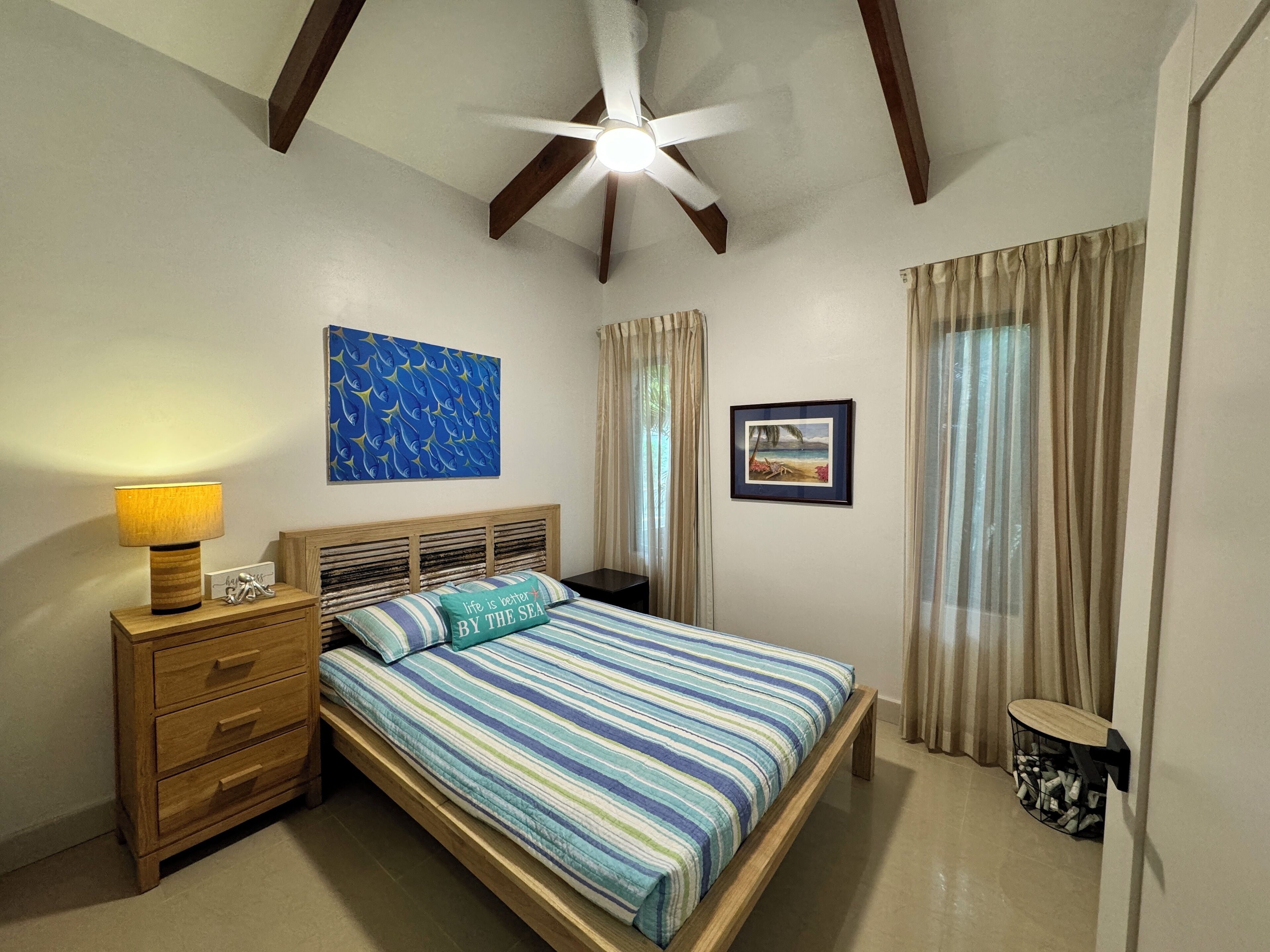 Une chambre à coucher comprenant un lit, une table de chevet, une lampe, un ventilateur de plafond, une fenêtre avec des rideaux et des œuvres d’art accrochées aux murs.