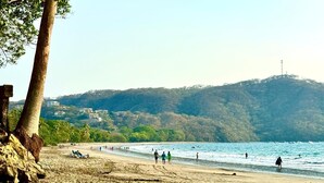 Ein Strand mit Menschen, die spazieren gehen und schwimmen, eine felsige Küste und Gebäude im Hintergrund.